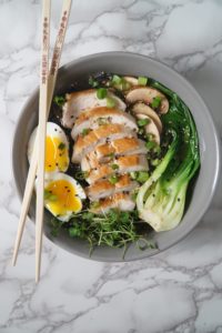 A grey bowl with chopsticks, broth, black noodles, bok choy, chicken, egg,s and greens for a Asian Chicken noodle soup bowl 