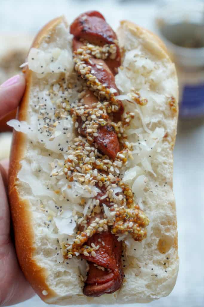 Holding a spiralized hot dog in a bun topped with sauerkraut, spicy mustard, and everything bagel seasoning. 